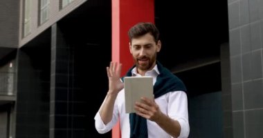 Gülümseyen sakallı işadamı dijital tablet üzerine video konferansında konuşuyor ve el kol hareketi yapıyor. Çevrimiçi iletişim sırasında şehir merkezinde duran genç adam.