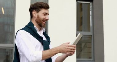 Açık hava konferansı için dijital tablet kullanan beyaz tişörtlü kendine güvenen sakallı bir adam. İş adamı ofis dışında online toplantı yapıyor..