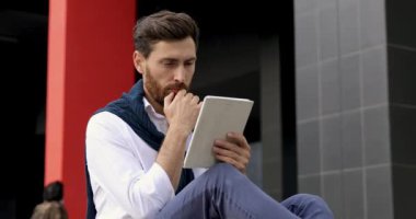 Beyaz gömlekli bir iş adamı iş meselelerini çözmek için dijital tablet kullanıyor. Sakallı adam temiz havada oturuyor ve modern aletlere bakıyor..