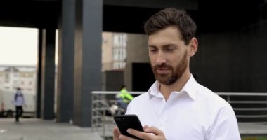 Kendine güveni olan sakallı bir adam ofis merkezinin yanında dururken modern akıllı telefondan internete göz atıyor. Mutlu adam temiz hava almak için işe ara veriyor..