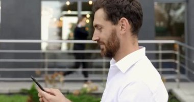 Gülümseyen sakallı bir adamın modern iş merkezinin yakınında yürüdüğünü ve elinde akıllı telefon tuttuğunu görüyorum. Beyaz gömlekli mutlu işadamı ofis dışındaki e-postaları kontrol ediyor..