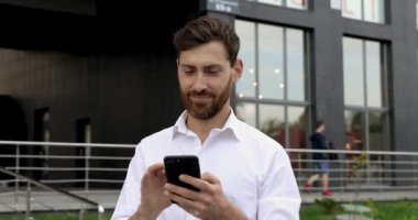 Sakallı ve saç kesimli gülümseyen adam akıllı telefon ekranında işaret parmağıyla oynuyor. Sokakta dikilirken modern aletler kullanan beyaz gömlekli yakışıklı bir adam..