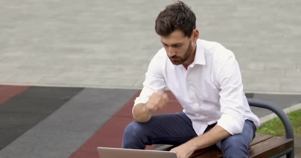 Bedachtzame man met een baard die op een bank zit met een geopende draadloze laptop en op afstand werkt. Knappe man met wit shirt en blauwe jeans. — Stockvideo