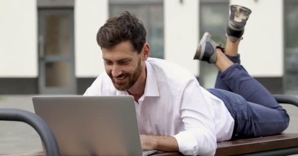 Hombre feliz con corte de pelo de moda y barba acostado en el banco y trabajando en el ordenador portátil inalámbrico durante los días cálidos al aire libre. Freelancer masculino disfrutando de su trabajo favorito al aire libre. — Vídeos de Stock