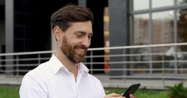 Veselý muž s přesnými vousy a účesem chodí po ulici a používá smartphone. Fešák v bílé košili procházení internetu na čerstvém vzduchu. — Stock video