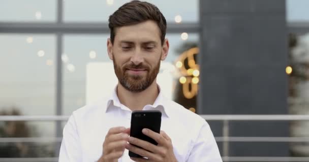 Портрет успешного бизнесмена в белой рубашке, улыбающегося и смотрящего в камеру, стоя на улице со смартфоном. Бородатый парень использует современный гаджет для работы на открытом воздухе. — стоковое видео