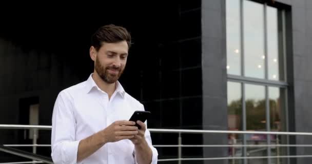 手にスマートフォンでオフィスセンターの近くを歩く白いシャツを着た快適な髭の男の肖像画。モバイル屋外での幸せなビジネスマンのテキストメッセージ. — ストック動画