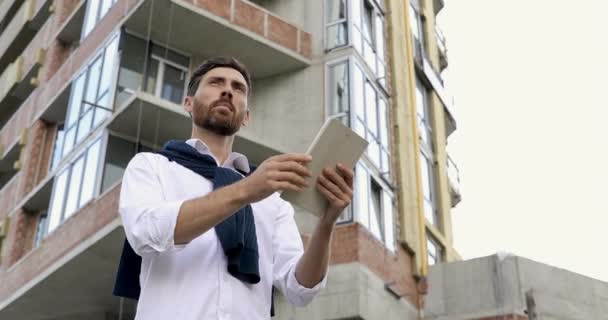 Hombre barbudo serio en camisa blanca comprobando realizado proyecto de construcción. Ingeniero experimentado usando gadget digital para el trabajo en el aire fresco. — Vídeos de Stock