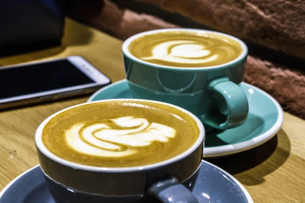 Fresh coffee in coffee shop. Latte - an art on a cappuccino. Tasty and beautifully. Two cups on a table. Phone. Drawing swan. — Stock Photo, Image