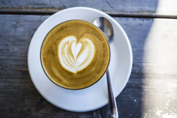 Copo de vidro grosso com cappuccino e latte-art no coração forma. Café fresco com leite. Em uma mesa de madeira com pires, colher — Fotografia de Stock