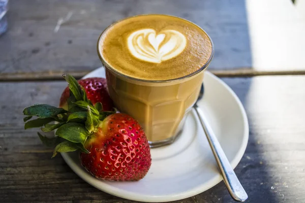 Thick glass tumbler with cappuccino and latte-art in the form heart. Fresh strawberry and coffee. On wood with saucer, spoon.