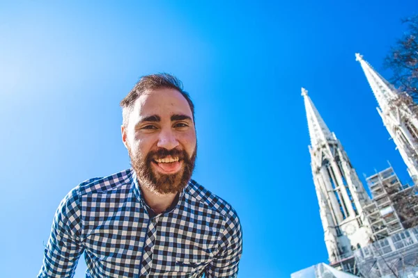 O jovem barbudo de camisa sorri contra o fundo do céu azul e as torres da catedral de Viena. Positivo e feliz . — Fotografia de Stock