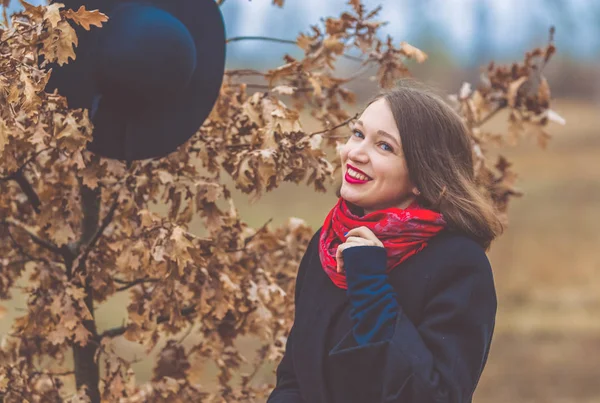 Genç Kız parkta bir siyah ceket sonbaharda yürür, şapka bir ağaç üzerinde asılı. Kırmızı ruj. Moda ve stil.