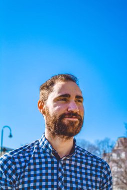 Portre genç adam parkta açık mavi gökyüzünün arka planda gömlekli duygularla sakallı. Sokak moda, yenilikçi.