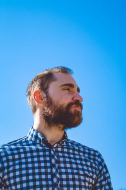 Portre genç adam parkta açık mavi gökyüzünün arka planda gömlekli duygularla sakallı. Sokak moda, yenilikçi.