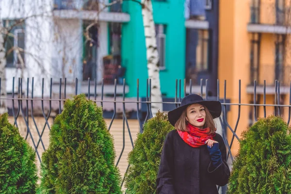 A jovem caminha na rua da cidade contra o fundo de edifícios multicoloridos em casaco preto e chapéu. Moda e estilo — Fotografia de Stock