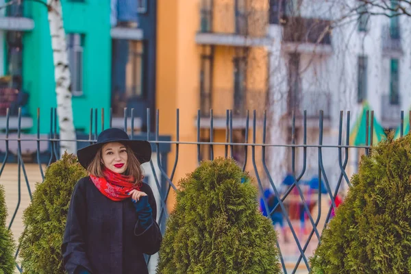 A jovem caminha na rua da cidade contra o fundo de edifícios multicoloridos em casaco preto e chapéu. Moda e estilo — Fotografia de Stock