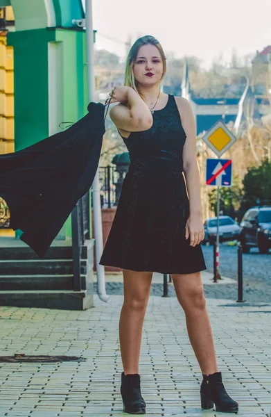 La muchacha joven en la ropa negra en el día soleado primaveral pasea por las calles viejas de la ciudad. Vestido, chaqueta y abrigo. Moda moderna . — Foto de Stock