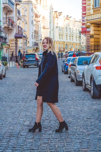 Kiev, Ucrânia. 30.03.2019. A menina jovem na roupa preta no dia ensolarado de primavera anda em ruas de cidade velhas. Vestido, casaco e casaco . — Fotografia de Stock