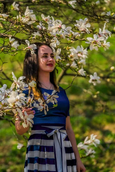 Молодая девушка в длинном платье гуляет в весеннем парке среди цветущих деревьев белых магнолий. Солнечная и ветреная погода. Юбка Макси . — стоковое фото
