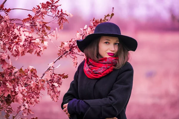Молодая девушка гуляет по осеннему парку среди деревьев в черном пальто и шляпе. Красная помада. Мода и стиль. Ноябрь . — стоковое фото