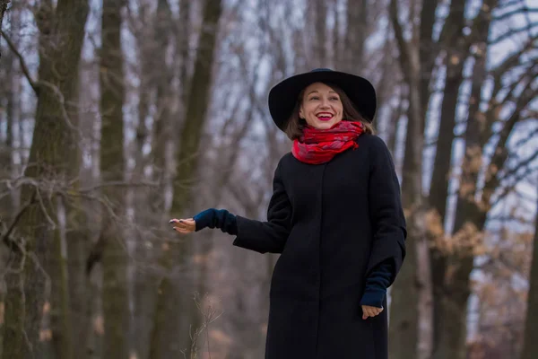 La ragazza in un soprabito nero, un cappello di pala e con rossetto rosso su labbra, cammina nel parco. Stile e moda moderna . — Foto Stock