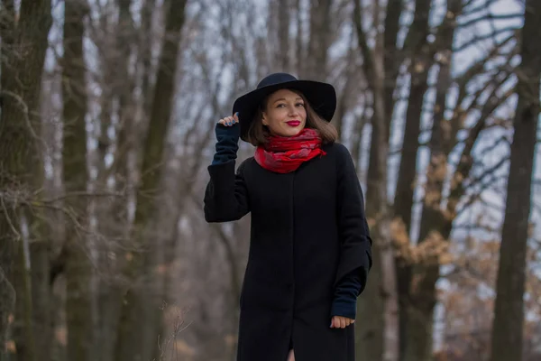A menina com um casaco preto, um chapéu de pá e com batom vermelho nos lábios, caminha no parque. Estilo e moda moderna . — Fotografia de Stock