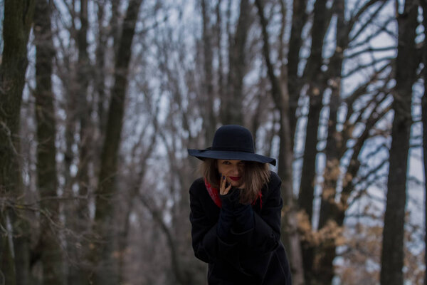 The girl in a black coat, a shovel hat and with red lipstick on lips, walks in the park. Style and modern fashion