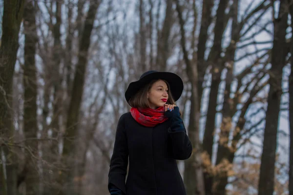 La chica con un abrigo negro, un sombrero de pala y con lápiz labial rojo en los labios, camina por el parque. Estilo y moda moderna . — Foto de Stock