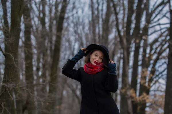 La jeune fille en manteau noir, un chapeau de pelle et avec du rouge à lèvres sur les lèvres, se promène dans le parc. Style et mode moderne . — Photo