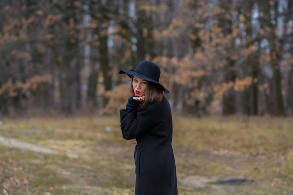 Dziewczyna w czarnym płaszczu, łopata i kapelusz z czerwoną szminką na ustach, spacery w parku. Styl i Nowoczesna moda. — Zdjęcie stockowe