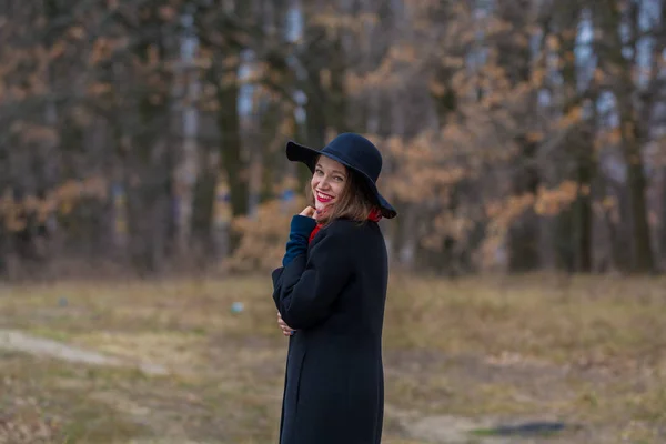 La chica con un abrigo negro, un sombrero de pala y con lápiz labial rojo en los labios, camina por el parque. Estilo y moda moderna . — Foto de Stock