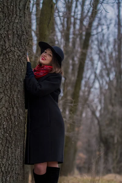 La chica con un abrigo negro, un sombrero de pala y con lápiz labial rojo en los labios, camina por el parque. Estilo y moda moderna . — Foto de Stock