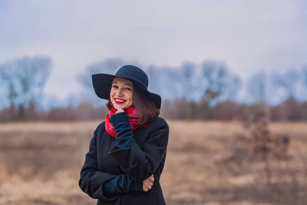 La chica con un abrigo negro, un sombrero de pala y con lápiz labial rojo en los labios, camina por el parque. Estilo y moda moderna . — Foto de Stock