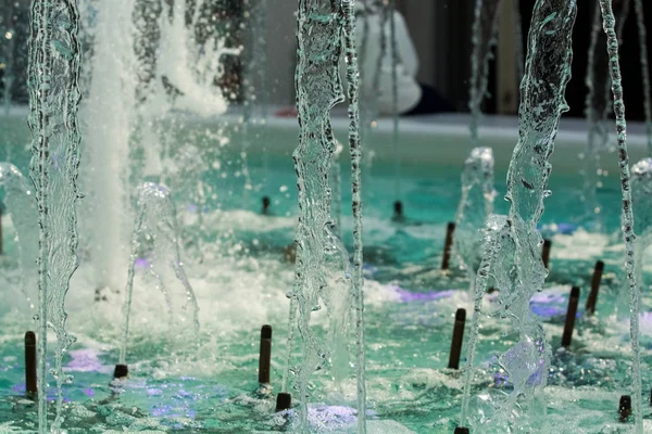 Big and strong water jets from the fountain with blue illumination in shopping center. Splashes and drops.