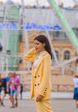 Genç ve şık kız sarı clasic takım elbise li dönme dolap karşısında şehir etrafında yürüyor. Moda.