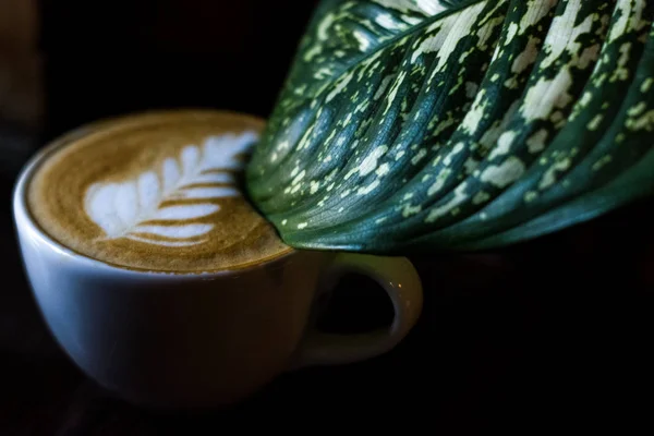 卡布奇诺在白色陶瓷杯与拿铁艺术和大绿叶在杯子从花 在黑色背景的咖啡店中喝的饮料 — 图库照片