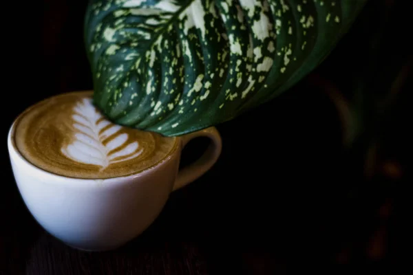 ラテアートと花からカップに大きな緑の葉と白いセラミックカップのカプチーノ 黒い背景のコーヒーショップで香りの良い飲み物 — ストック写真