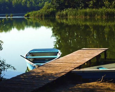 Nehirde yaz sabahı, rıhtımda tekne suya, ormana, çimenlere yansıyor.