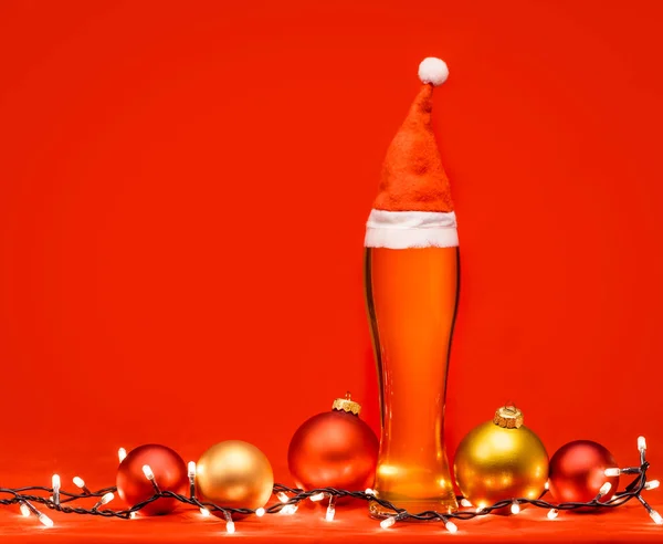 特写镜头全皮斯纳杯苍白的啤酒或啤酒与圣诞老人或圣诞节红色帽子 圣诞灯和在红色背景上的小球 — 图库照片