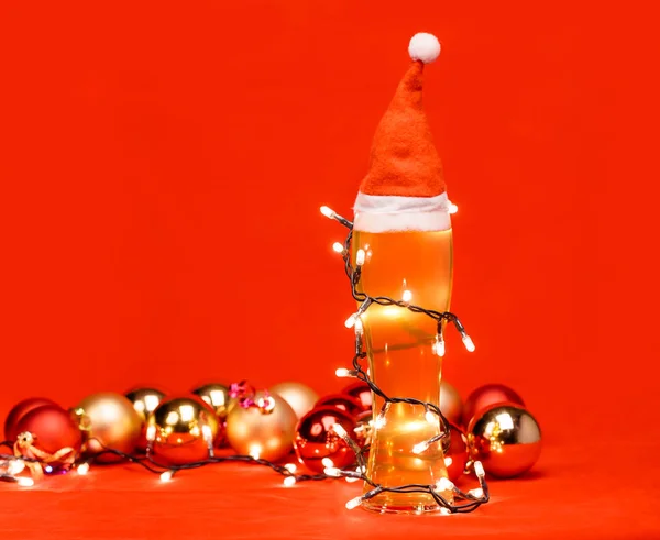 特写镜头的苍白啤酒或啤酒与圣诞老人或圣诞红帽的顶部 包裹在圣诞灯和圣诞小球背景下的圣诞泡泡 — 图库照片