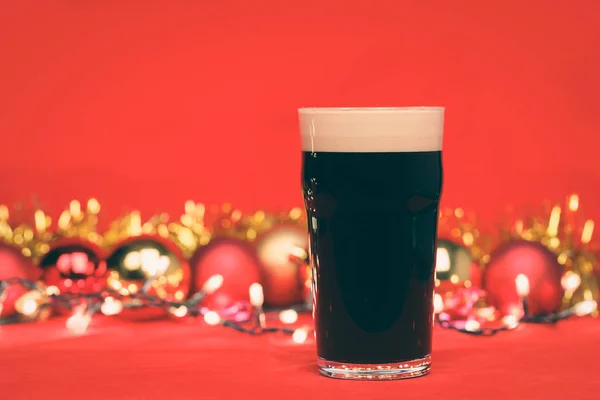 Nonik pint glass of dark beer or stout ale with christmas lights baubles and tinsel on red background