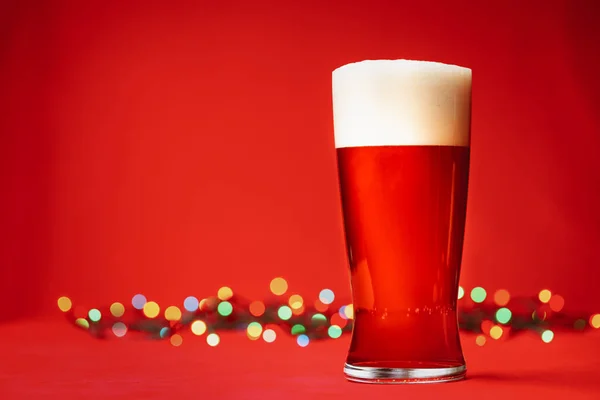 Pint glass of of pale lager beer or ale with big head of foam and christmas lights on red background