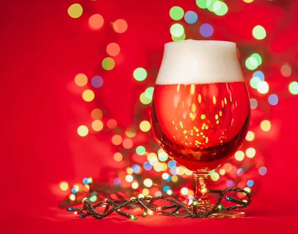 Snifter glass of pale lager beer or ale with  christmas lights on red background