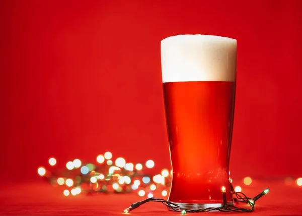 Pint glass of of pale lager beer or ale with big head of foam and christmas lights on red background