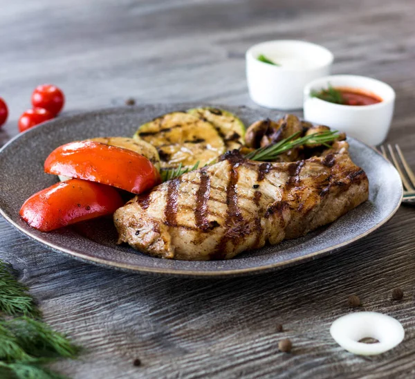 Grilled loin and grilled vegetables are on a plate in the kitchen. Grilled piece of meat with strips and vegetables on a wooden gray table