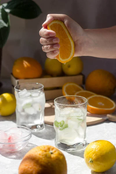 A glass filled with ice. From above, a woman\'s hand squeezes drops of orange juice into a glass