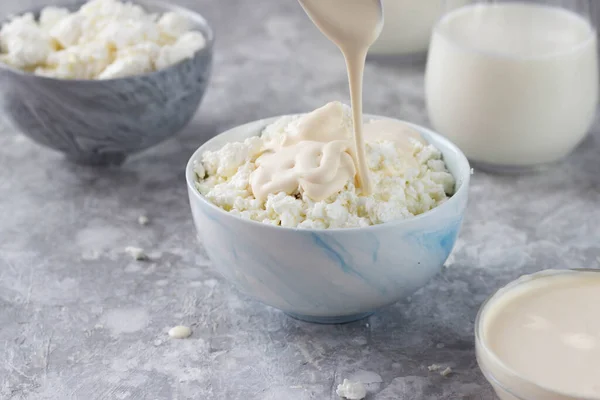 Large chunks of cottage cheese lie in a plate, and sour cream pours from a spoon on top of them. Glasses of milk are at the back