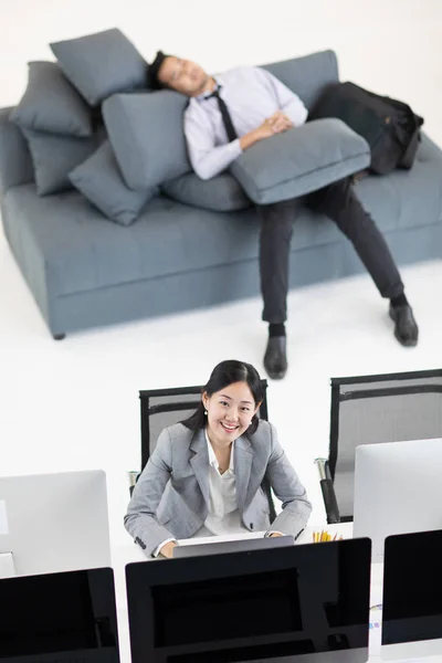 Businessman sleeping on sofa in working hours while business woman working and typing computer at desk, concept for time cheating in office