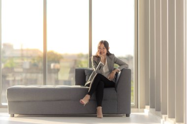 Bir uzun saçlı Asyalı bağdaş kurup mutlu arkadaki büyük pencereler ile odada gülümseyen kanepede oturuyor.